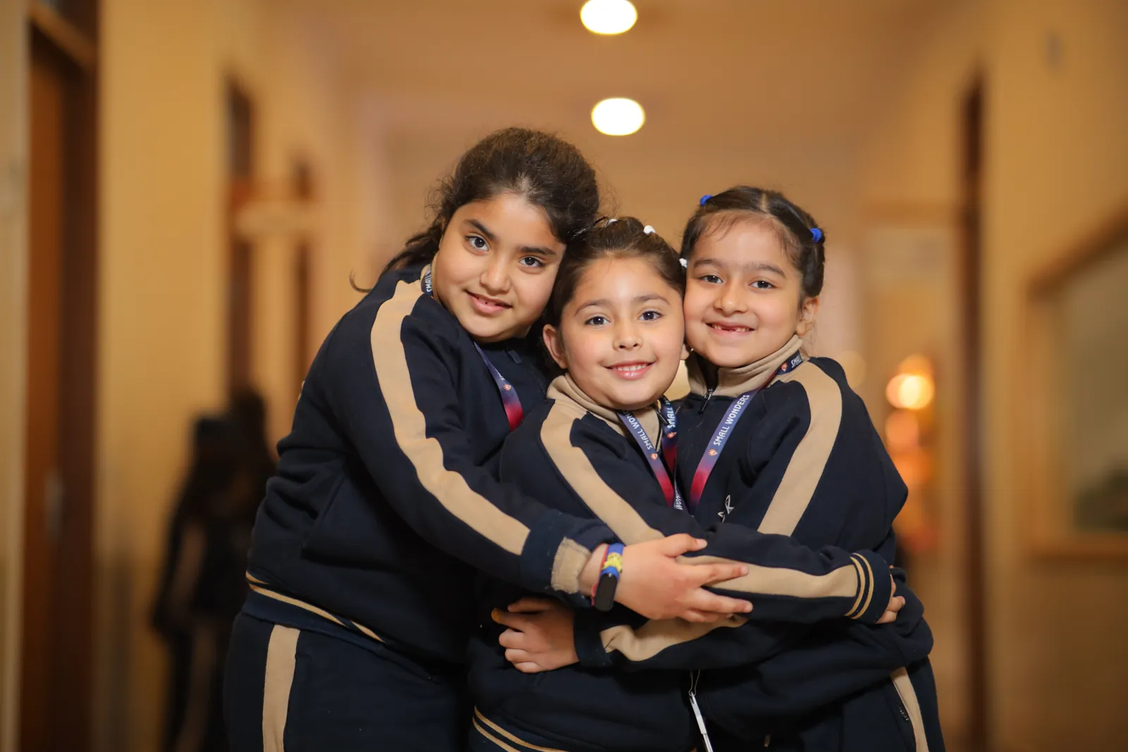 Three learners hugging and smiling in a school corridor.